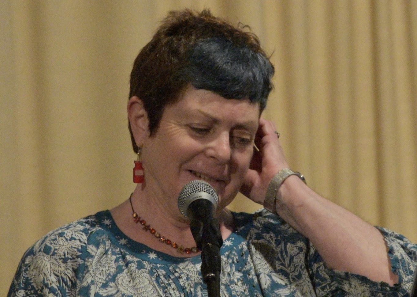 Head and Shoulder image of Pat Edwards talking into a microphone. She is looking down slightly to her left. She wears a blue top with a white flowery pattern .
