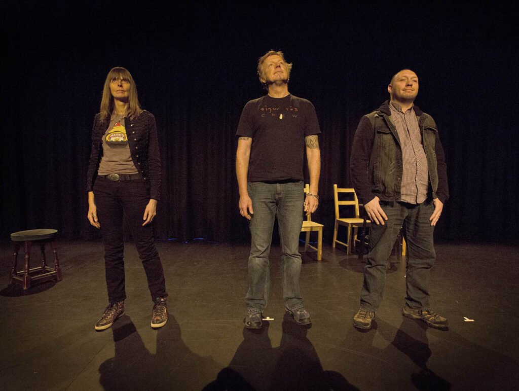 The three P's stand in a line. Emma is on the left, Steve in the middle, Dave on the right. They are staring into the air. The shot is taken on a stage. Three chairs and a stool can just about be seen in the background.