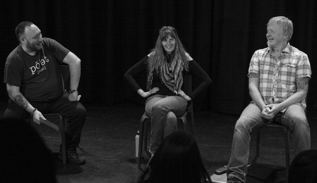 The three P's sit in a line. Dave on the left, Emma in the middle and Steve on the right. They are all smiling. Dave has one hand on his knee and is looking at Steve who is looking straight back. Emma looks out into the audience.