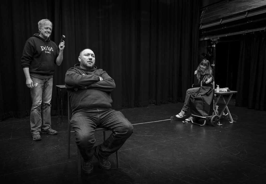 A black and white still from the show Whose Round Is It Anyway. Dave sits at the front, arms folded, looking into space. To his left and slightly back is Emma sitting cross legged and smiling. Behind Dave is Steve, a mischevious grin on his face as he holds a pair of hairclippers in his left hand.