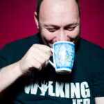 Dave sits in front of a deep red background. He wears a black T-Shirt which reads "The Walking Jed" and is laughing while leaning forward and drinking from a blue and white mug.