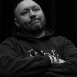Dave sits leaning back, arms folded. The image is black and white and he wears a dark hoodie with the word "Poet" on it. On his right hand are two bracelets. He has a thoughtfull look on his face as he stares into the distance.